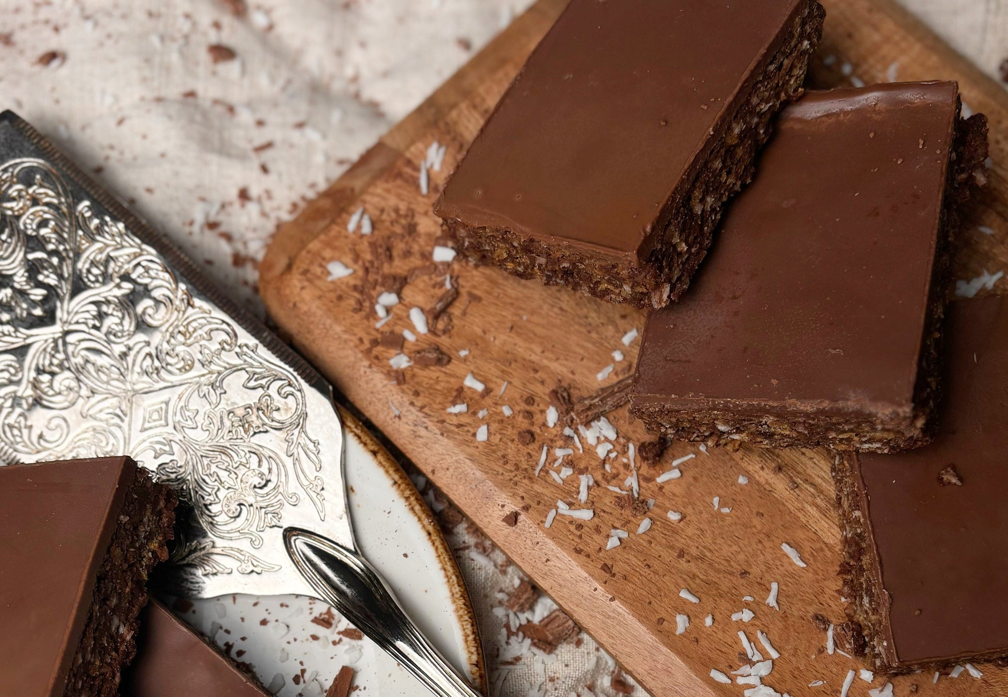 Coconut crunch slices topped with chocolate on a wooden board with a decorative silver knife and spoon.